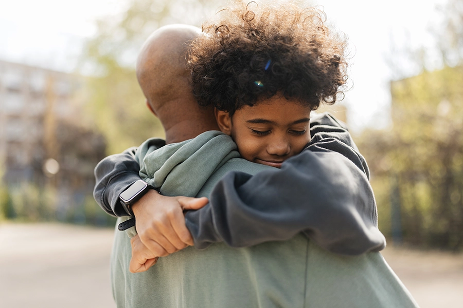 Young boy with curly hair hugging an adult tightly outdoors, eyes closed and smiling.